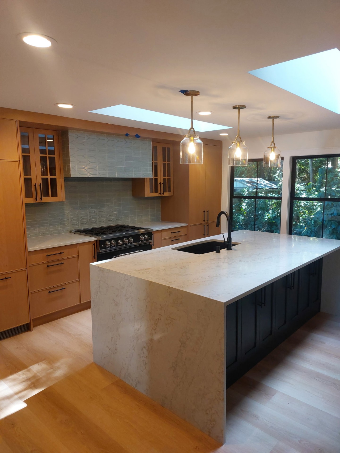 Modern kitchen with quartz waterfall island, warm wood cabinets, black island base, brass pendants and skylights